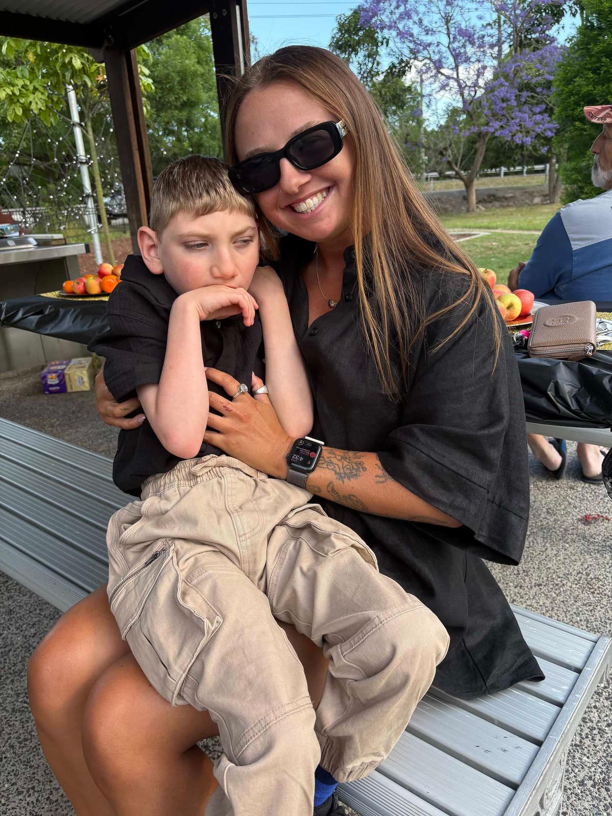 A Pal Supports worker sitting with a young boy on her lap at a park in the Hunter, jacarandas in flower behind them